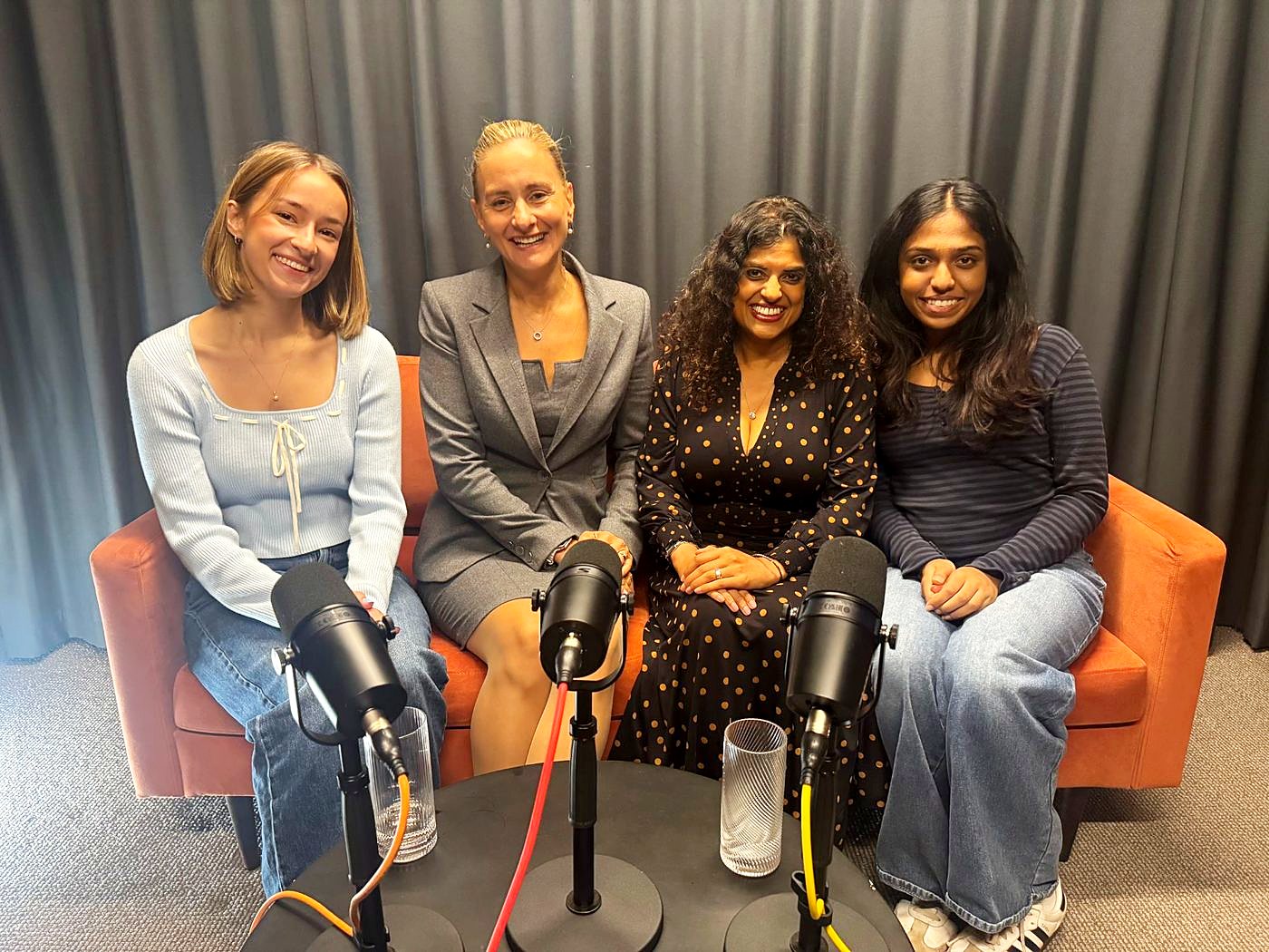 Four guests smiling together on an orange sofa behind three microphones in The Dokkcast studio